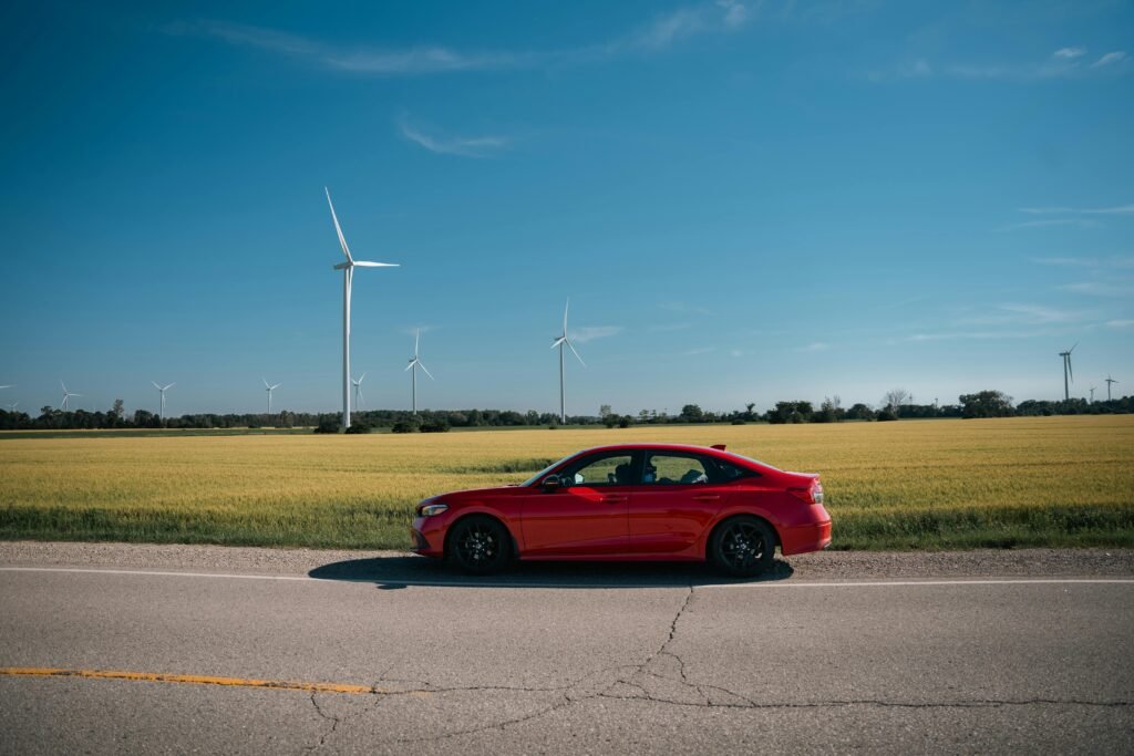 A red sedan on a rural road beside fields with wind turbines under a clear blue sky.